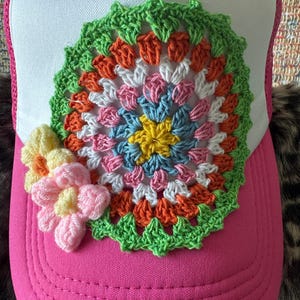 May include: A bright pink trucker hat with a white front panel. The hat features a large, colorful, crocheted floral design in shades of yellow, blue, pink, orange, and green. A smaller crocheted flower in pastel colors is on the side.