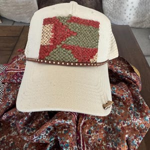 May include: Cream-colored trucker hat with a woven patch featuring red, green, and beige geometric shapes. The hat has a brown leather band with gold studs and a small gold-colored metal accent on the brim. The hat is resting on a floral patterned scarf.