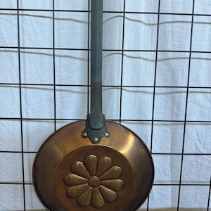May include: A round copper pan featuring a central embossed flower design with twelve petals. The pan has a long, dark gray metal handle attached with a three-point bracket secured by three visible copper rivets at the base of the handle.