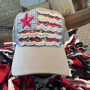 Puede incluir: Una gorra de béisbol gris claro con un panel frontal decorativo que presenta un cuadrado de cuadros azules y blancos, una sección de tela floral rosa, una estrella bordada rosa brillante y filas horizontales de encaje blanco festoneado.