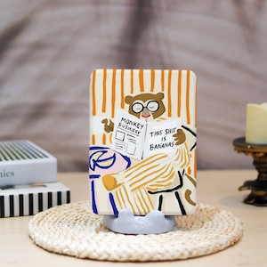 May include: A rectangular phone stand with a cartoon monkey reading a newspaper titled "Monkey Business" and "This Shit Is Bananas." The stand has a beige background with orange vertical stripes. The monkey wears glasses and is in pajamas.