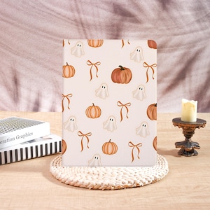 May include: A rectangular tablet case with a light cream background featuring a repeating pattern of orange pumpkins, white ghosts, and brown ribbons. The case is positioned upright on a round woven mat, displaying a festive seasonal design.