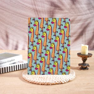 Könnte beinhalten: Ein Notizbuch mit einem sich wiederholenden Muster aus gelben, orangefarbenen und rosa Cartoon-Giraffen auf hellblauem Hintergrund. Die Giraffen tragen Brillen und Schuhe. Das Notizbuch liegt auf einem gewebten Platzset.