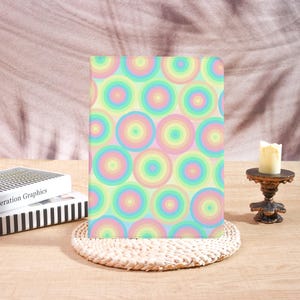 May include: A pastel-colored notebook with a pattern of concentric circles in shades of pink, yellow, blue, and green. The notebook is resting on a woven placemat. A candle sits on a decorative stand.