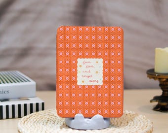 May include: An orange notebook with a floral pattern and the words "fun, sun, and target ruins" on a white label. The notebook is in a grey stand on a woven mat.