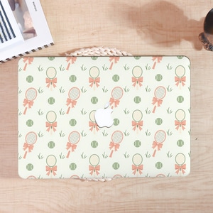 May include: A laptop case with a tennis theme. The case features a repeating pattern of tennis rackets with pink bows, green tennis balls, and blades of grass on a cream background. The Apple logo is visible.