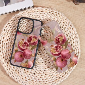 May include: Two phone cases with a pomegranate design. The cases feature a black border and a clear back with a pattern of pink pomegranates, green leaves, and gold accents. The design is set against a light purple background.