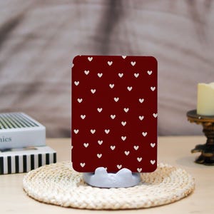 Könnte beinhalten: Ein rechteckiger, weinroter Gegenstand mit einem Muster aus kleinen, weißen Herzen. Der Gegenstand wird von einem weißen, dekorativen Ständer gehalten. Der Hintergrund umfasst eine gewebte Matte, Bücher und einen Kerzenhalter.