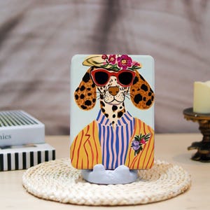 May include: A decorative phone case featuring a Dalmatian dog wearing sunglasses, a hat with flowers, and a striped shirt. The dog has black spots and is set against a light blue background. The case is on a white stand.