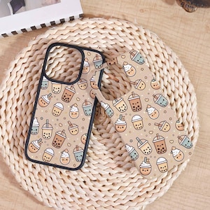 May include: Two phone cases with a repeating pattern of cartoon bubble tea drinks in various colors, including brown, blue, and white, on a tan background. One case is on top of the other, with a black border. The cases are on a woven, beige surface.