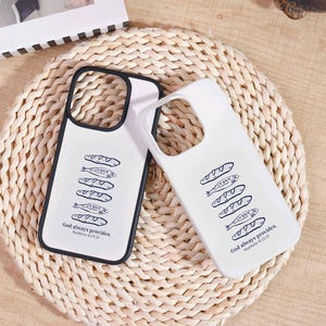 May include: Two white phone cases with a black outline. Each case features a black illustration of bread and fish, with the text "God always provides. Matthew 14:13-21." The cases are on a woven, beige surface.
