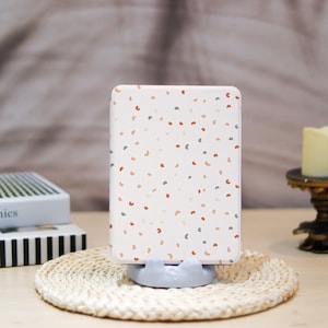 May include: A white rectangular phone case with a pattern of small, colorful, semi-circle shapes. The case is propped up on a light blue phone stand, resting on a woven coaster. The background includes books and a candle.