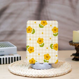 May include: A rectangular phone case with a yellow floral pattern on a light yellow and white checkered background. The case is resting on a light gray phone stand, and a woven coaster is beneath it.