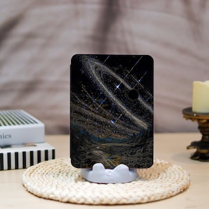 Peut inclure: Une coque de tablette noire avec un motif cosmique de spirales blanches et dorées, d'étoiles et d'un fond sombre. L'étui est soutenu par un support blanc, posé sur un tapis tissé. Une bougie et des livres sont en arrière-plan.
