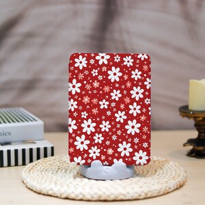 May include: A rectangular phone case featuring a red background with a pattern of white and pink flowers. The case is held by a light gray stand on a woven mat. A candle and books are in the background.
