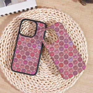 May include: Two phone cases with a repeating pattern of smiley faces with heart-shaped eyes in shades of pink, red, and purple. The cases have a black border and are set against a woven background.