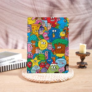 May include: A rectangular notebook featuring a vibrant, multi-colored doodle pattern filled with various cartoon characters, including a yellow smiley face, a slice of pizza, a green frog, and a tree, all set against a bright blue background.