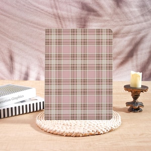 May include: A pink and brown plaid notebook stands upright on a woven coaster. A small candle sits on a decorative stand to the right. A book with the text "eration Graphics" is visible to the left.