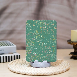 May include: A teal phone stand featuring a speckled pattern in pink, gold, and white. It sits on a woven coaster. A white book and a candle are visible in the background.