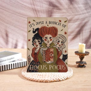 May include: A rectangular sign featuring an illustration of three women with distinct hairstyles and a black cat. The text reads IT'S JUST A BUNCH OF HOCUS POCUS. The central figure holds a brown book with a central eye design, surrounded by autumn leaves.