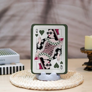 May include: A playing card featuring the Queen of Hearts, depicted with sunglasses, a cigarette, and a glass of wine. The card has a green border and is displayed on a white stand. The card is set on a woven mat.