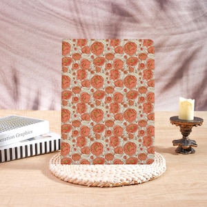 May include: A floral patterned notebook featuring a repeating design of orange and gold peonies on a cream background. The notebook is resting on a woven placemat, with a candle and books in the background.