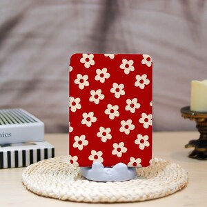 May include: A rectangular object with a red background and a repeating pattern of white flowers. The object is held in a light gray stand and rests on a woven mat. A candle and books are in the background.
