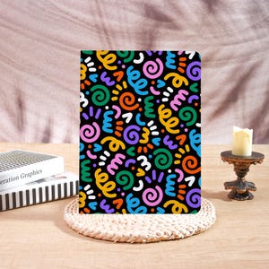 May include: A colorful notebook with a black background and a pattern of swirls, squiggles, and dots in yellow, orange, blue, green, purple, and white. The notebook is on a woven placemat.