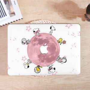 May include: A white laptop skin featuring a large pink moon at the center with a cutout for an Apple logo. The design includes various illustrations of Snoopy and Woodstock characters arranged around the moon, accented by small pink stars and dots.