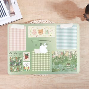 May include: A green laptop skin with various decorative stickers. The stickers include gingham patterns in pink and blue, illustrations of animals, a strawberry, and a pond scene. The Apple logo is visible.
