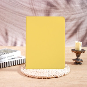 May include: A yellow tablet case on a woven placemat. A small candle sits on a decorative stand to the right. A book with the title "eration Graphics" is visible on the left. The background is a soft, textured fabric.