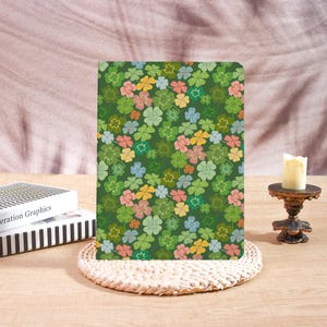 May include: A notebook with a green cover featuring a pattern of colorful four-leaf clovers in shades of green, pink, yellow, and blue. The notebook is resting on a woven placemat. A candle sits on a decorative stand.