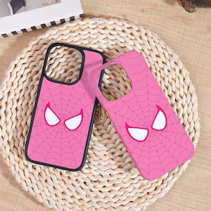 May include: Two pink phone cases with a black border. Each case features a spiderweb design and white eye shapes. The cases are on a woven, light brown surface.
