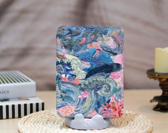 May include: A rectangular phone case featuring a vibrant underwater scene with marine life, including whales, jellyfish, and coral. The design incorporates shades of blue, pink, and green, creating a colorful and detailed illustration. The case is displayed on a white stand.