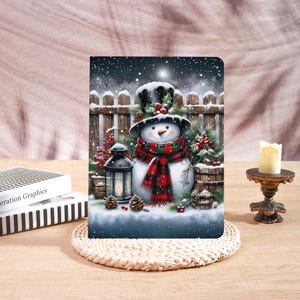 May include: A decorative notebook featuring a snowman in a winter scene. The snowman wears a black top hat and a red and green scarf. The background includes a wooden fence, snow, and a lantern. A lit candle sits on a small stand.