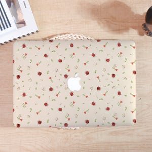 Peut inclure: Un ordinateur portable avec une coque beige ornée d'un motif répété de coccinelles rouges, de feuilles vertes et de coeurs rouges. Le logo Apple est visible au centre. L'ordinateur est sur une surface en bois clair.