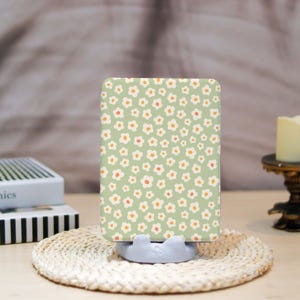 May include: A rectangular phone case with a floral pattern of white flowers with orange centers on a light green background. The case is propped up on a light gray phone stand, sitting on a woven coaster.