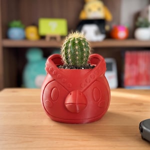 Puede incluir: Una maceta pequeña, redonda y roja con forma de cabeza de pájaro estilizada, con un pico prominente y marcas oculares circulares. Un cactus cilíndrico, verde y espinoso está plantado en el recipiente, situado sobre una superficie de madera clara.