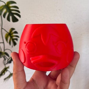 May include: A bright red, rounded planter featuring a molded, stylized face with large circular eyes, a small beak-like protrusion, and a leaf-shaped detail on the forehead. The container has a wide, open top and a smooth, matte finish.
