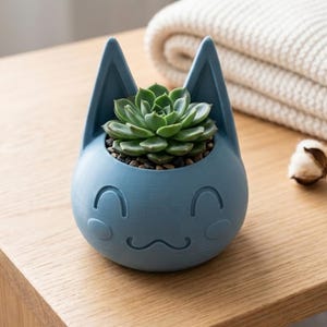 May include: A blue cat-shaped planter with a smiling face holds a green succulent plant. The planter has pointed ears and a round body. The succulent is surrounded by small pebbles. The planter sits on a wooden surface.