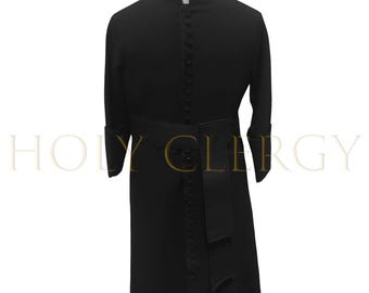 May include: A black clergy cassock with a mandarin collar and long sleeves. The garment features a button-down front, a belted waist, and decorative cuffs. The text "HOLY CLERGY" is visible in the background.