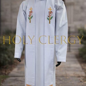 May include: A light blue clergy robe with long sleeves, featuring embroidered crosses and floral designs in gold, red, and green. The text "HOLY CLERGY" is displayed in gold. The robe has a high collar and a decorative gold trim.