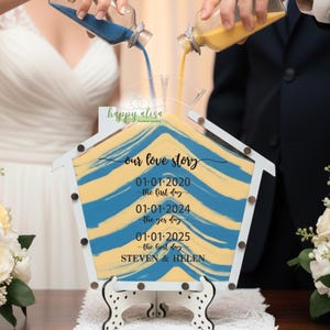 May include: A wedding sand ceremony set with a house-shaped wooden frame. The frame displays blue and yellow sand layers and the text "our love story" with dates. Two people pour sand from bottles into the frame.