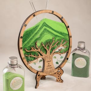 May include: A wooden sand ceremony set with a tree design. The set includes a round frame with green sand, a clear funnel, and two bottles of green sand. The frame has a tree cutout and the names "Steven & Helen 12.03.2026" are engraved.