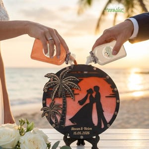 May include: A wedding sand ceremony set with a black wooden frame featuring palm trees and a silhouette of a couple. The set includes two bottles pouring sand into the frame, with the names "Steven & Helen" and the date "15.05.2026" engraved.