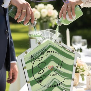 May include: A wedding sand ceremony set with a house-shaped container. Green sand is poured into the container, which has a green and white chevron design. The container is decorated with the initials "S & H" and the date "09.09.2024". The text "A beautiful unity" is at the top.