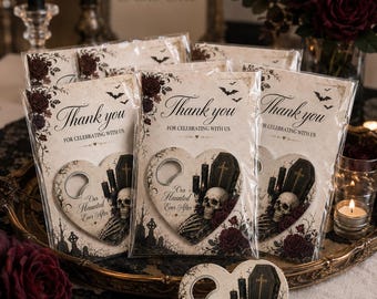 Può includere: Apribottiglie a forma di cuore con una stampa raffigurante un teschio, una bara, candele nere e rose rosso scuro. Il testo recita "Thank you FOR CELEBRATING WITH US" e "Our Haunted Ever After". Ogni articolo è sigillato in una busta di plastica trasparente.
