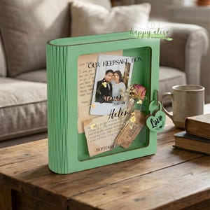 Può includere: Un portagioie verde menta a forma di libro, con un fronte trasparente che mostra una foto, note manoscritte e fiori secchi. La scatola è su un tavolo di legno. Il testo sulla scatola recita "OUR KEEPSAKE BOX" e "Steven and Helen".