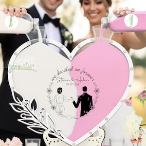 May include: A heart-shaped wedding sand ceremony set. The frame is white with a pink and white sand design. The text "we decided on forever" and the couple's names and wedding date are displayed. The couple is pouring sand.