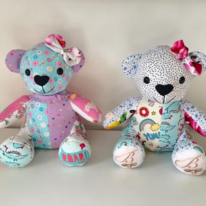 May include: Two handmade teddy bears with patterned fabric. One bear has a blue floral design, the other has a white polka dot pattern. Both have bows, heart-shaped noses, and embroidered details. The bears are sitting on a white surface.